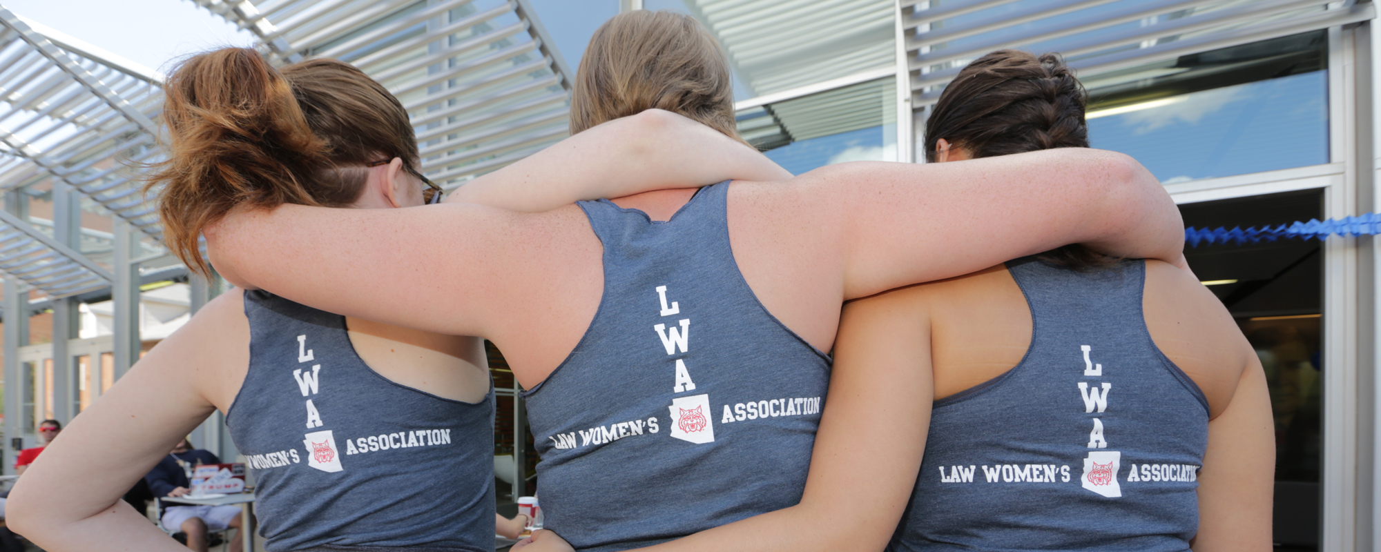 Three women link arms and display the backs of their shirts, promoting the Law Womens Association