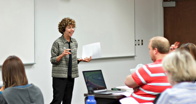 A woman, Barbara Atwood, stands at the front of a classroom, teaching a room of students.