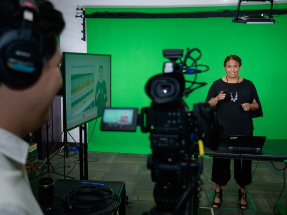A cameraman films a professor delivering a lecture in front of a green screen inside a recording studio