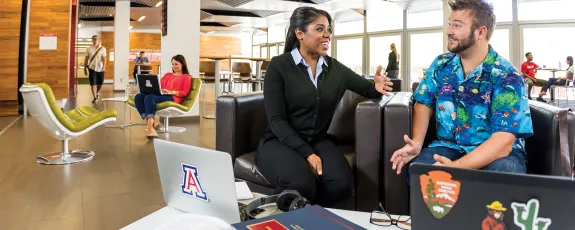 Two students sit on a couch in mid-conversation, surrounded by law textbooks, laptops and other students