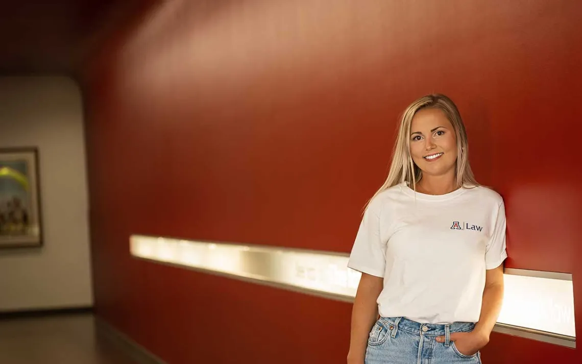 Student in white U of A tshirt standing against a bright red wall in the law building.